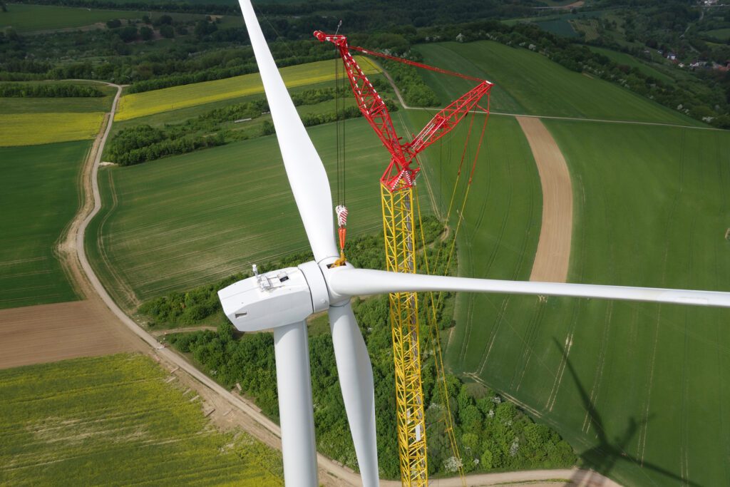 A large wind turbine is being assembled in a green, rural landscape using a tall yellow and red crane. Farmland and fields surround the construction site.