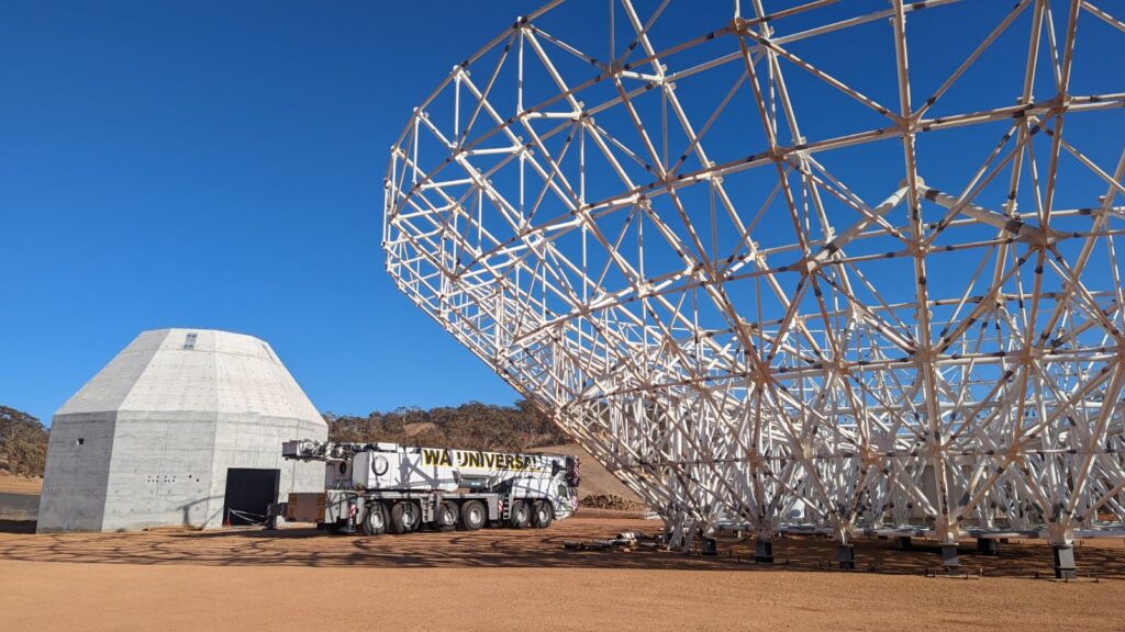 A large metal radio telescope dish under construction sits next to a concrete building and a truck labeled "WA UNIVERS" on a dirt surface, with clear blue sky and hills in the background.