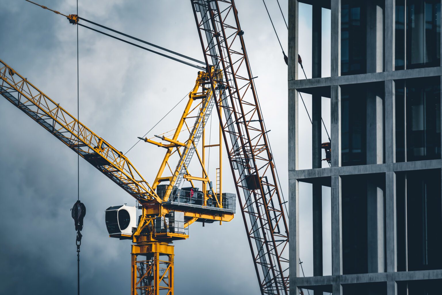 A close-up of two large construction cranes next to a modern building under construction, with a cloudy sky in the background. The cranes' yellow and red structures are prominent in the scene.