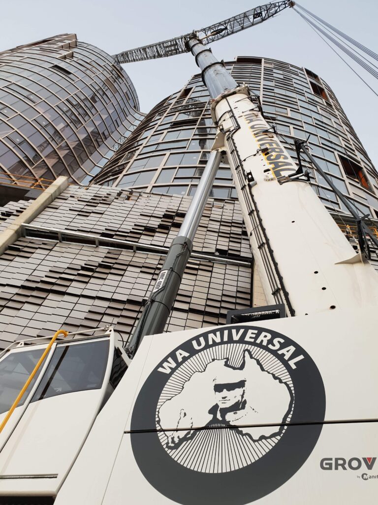 A large construction crane labeled "WA Universal" is positioned in front of modern, curved glass buildings, viewed from a low angle. The crane's arm extends upward, and construction appears to be ongoing.
