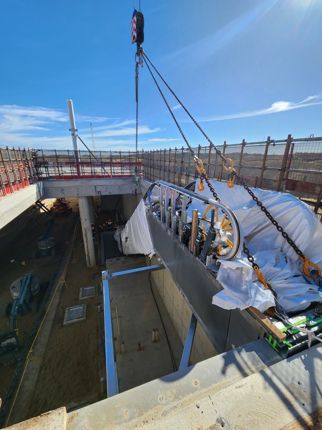 A large piece of machinery wrapped in white material is being lifted by a crane at a construction site, where workers and equipment are visible below near a partially built concrete structure.