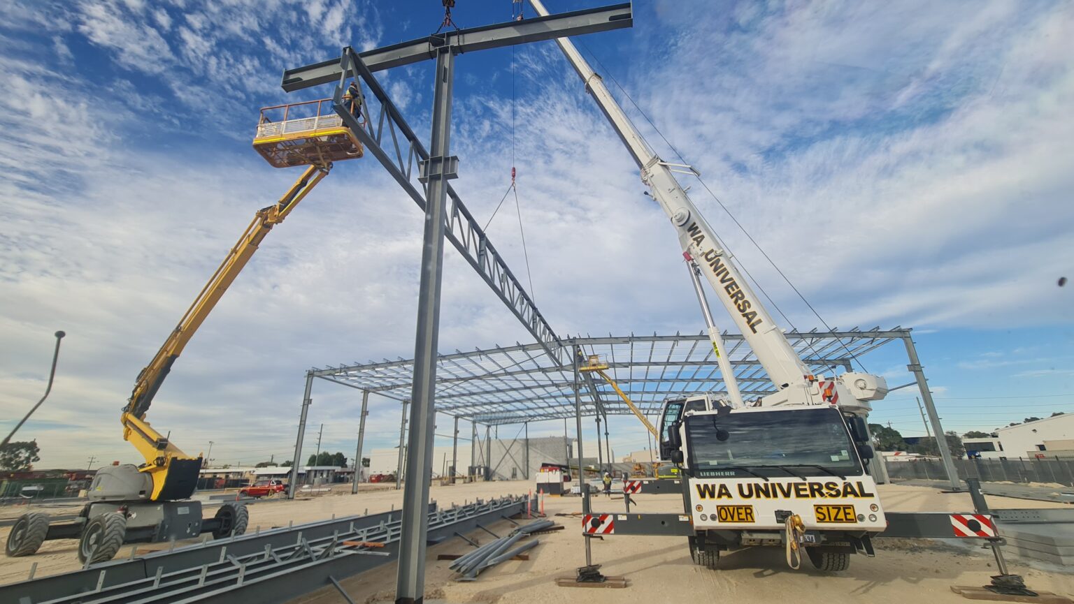 A crane and a boom lift are assembling a steel framework for a large industrial building. Workers are elevated, and materials are on the ground, with a bright blue sky in the background. The site appears under construction.