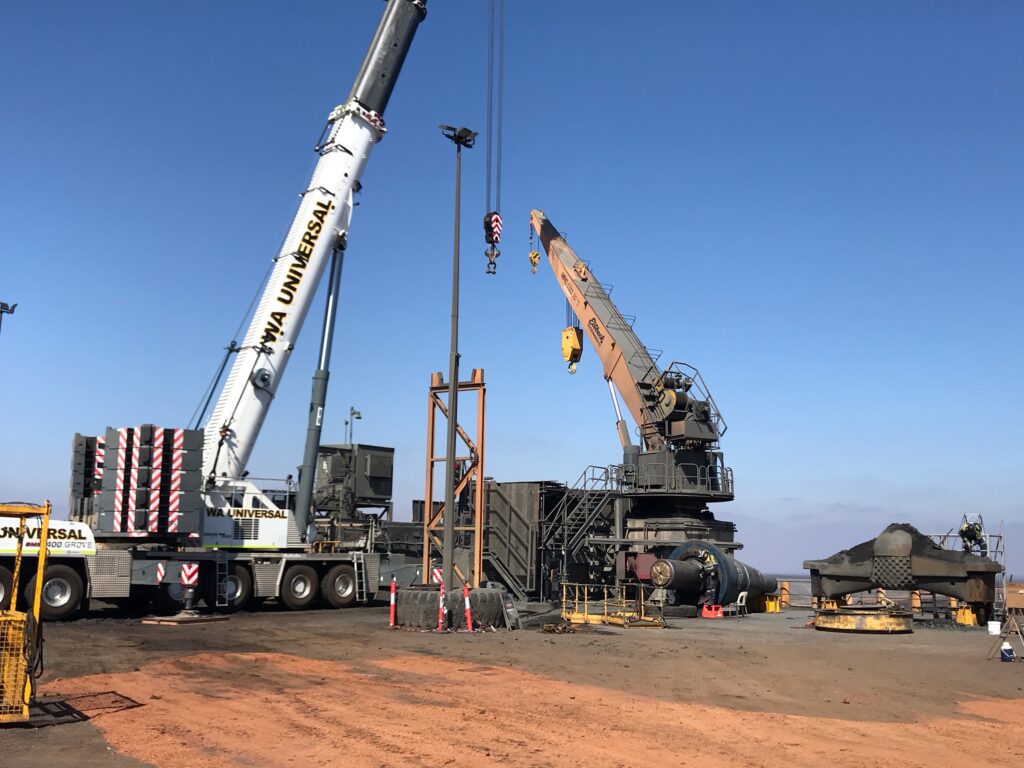 Two large cranes operate at an industrial mining site with machinery and equipment on reddish dirt under a clear blue sky. A few workers and safety barriers are visible in the area.
