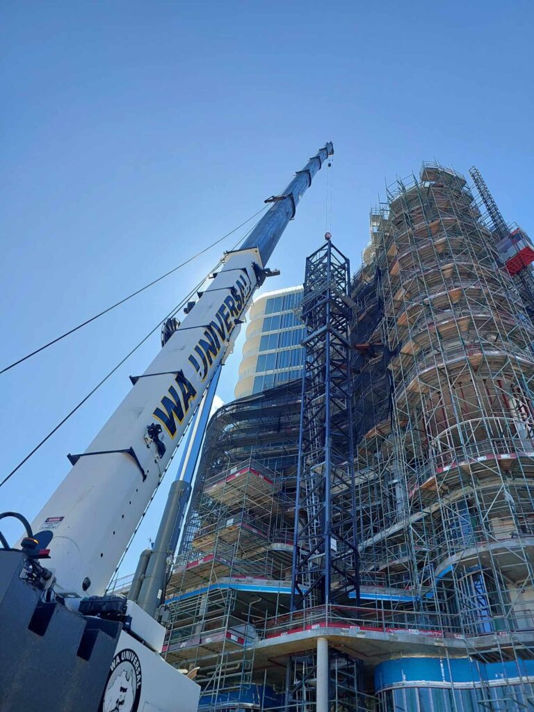 A large crane labeled "WA UNIVERSAL" lifts a metal structure beside a tall building covered in scaffolding under a clear blue sky, indicating active construction work.