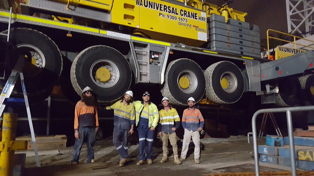 Five construction workers in high-visibility clothing and helmets stand in front of a large yellow crane at a worksite at night. The crane is raised off the ground, and the workers are posing and smiling.