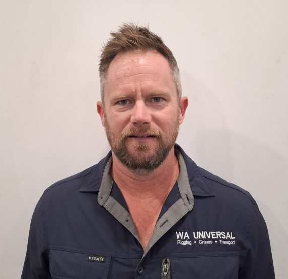 A man with short, light brown hair and a beard stands against a plain white wall, wearing a navy collared shirt with "WA UNIVERSAL Rigging • Cranes • Transport" embroidered on it.