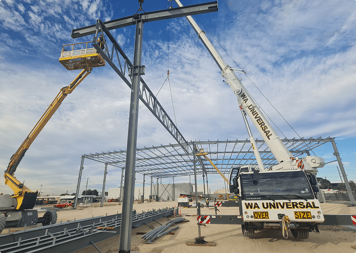 A construction site with workers on a yellow lift and a white crane labeled "WA UNIVERSAL" assembling a large steel frame structure under a blue sky with clouds.