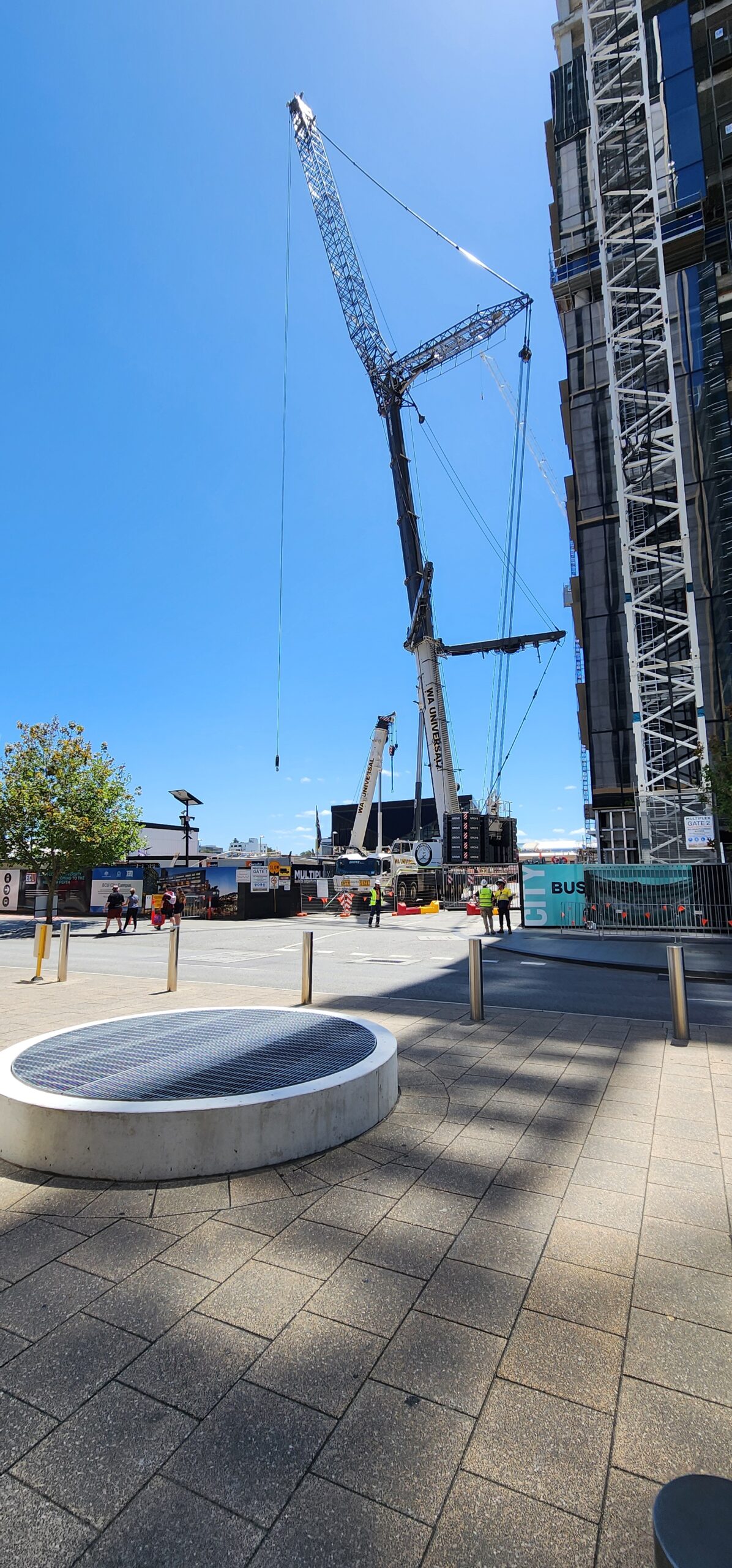 A large crane operates at a construction site next to a tall building under a clear blue sky. Several workers and construction barriers are visible in the foreground, along with pavement and a tree.