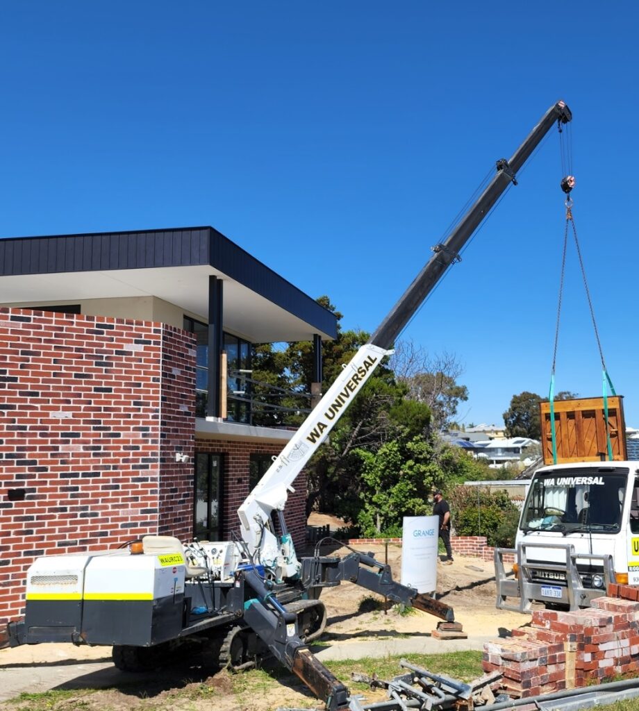 A small crane lifts a pallet of bricks from a flatbed truck outside a modern red-brick building under clear blue skies. Construction materials and equipment are scattered around the site.