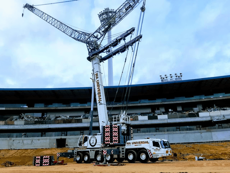 A large mobile crane labeled "WA Universal" is positioned on a sandy construction site in front of a partially demolished stadium with floodlights and a cloudy sky overhead.