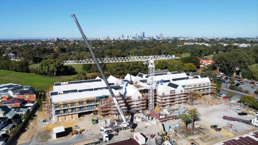 Aerial view of a large construction site with cranes and scaffolding around multiple buildings, surrounded by trees and houses, and a city skyline visible in the background under a clear sky.