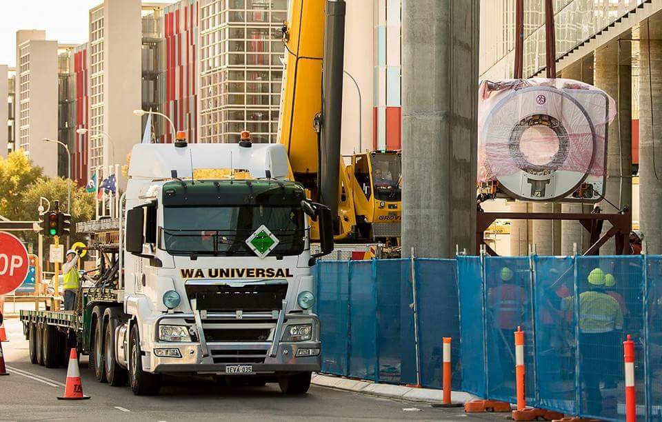 A truck labeled "WA UNIVERSAL" delivers large machinery as workers and a crane help unload it near a construction site, with blue fencing and traffic cones along the street.