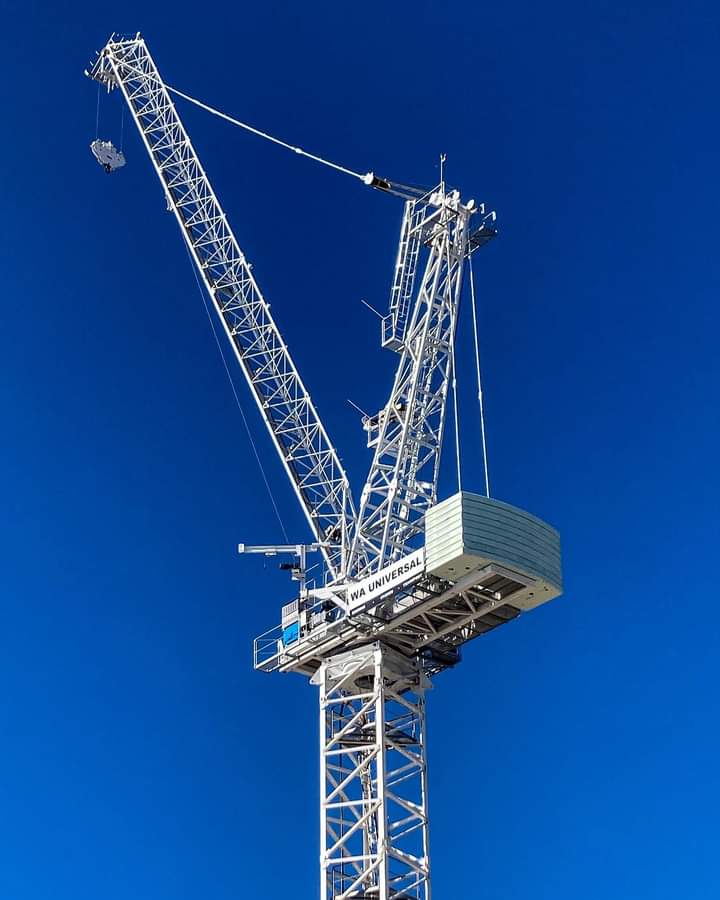 A tall white construction crane extends into a clear blue sky. The crane's arm and counterweight are visible, with the name "WA Universal" displayed on its side.