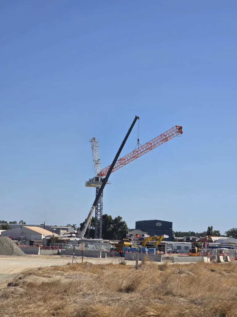 A construction site with a tall crane lifting materials, surrounded by machinery and buildings under a clear blue sky. Dry grass and some dirt piles are visible in the foreground.