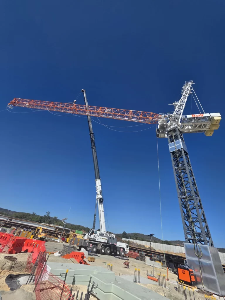 A large crane is lifting the arm of a tower crane at a construction site under a clear blue sky. Construction materials and safety barriers are scattered around the site.