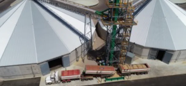 Aerial view of two large, round grain silos with metal roofs, a conveyor system between them, and two trucks parked beneath the conveyor loading or unloading grain.