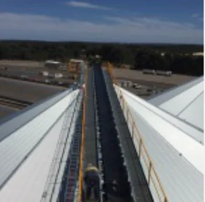A wide conveyor belt transports black material between large industrial buildings, with trees and parked vehicles visible in the background under a blue sky.