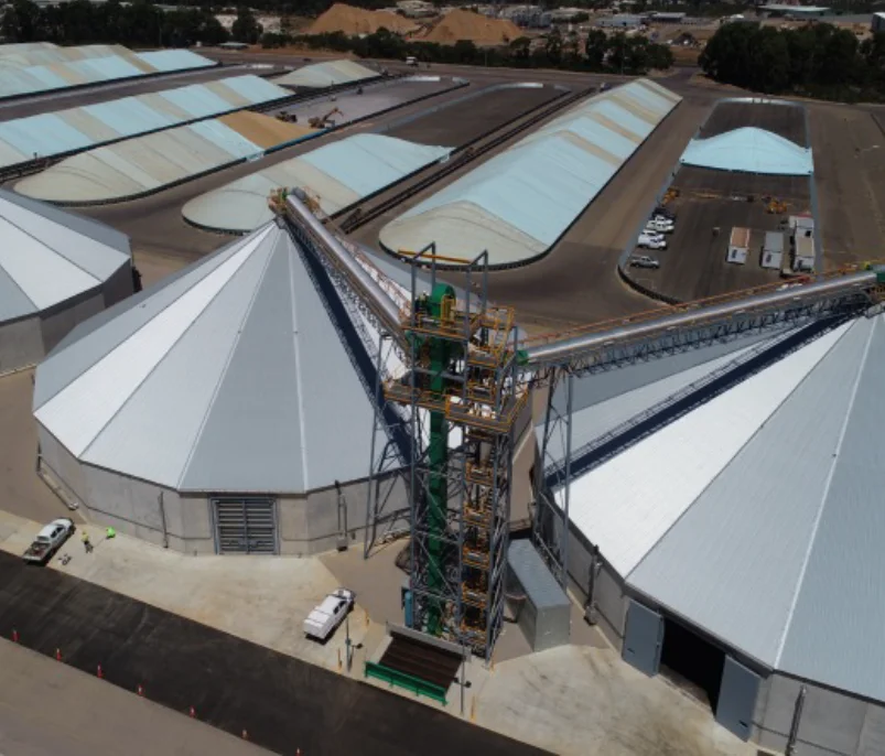 Aerial view of large conical and rectangular industrial storage buildings with light-colored roofs, connected by elevated conveyor belts, in a sprawling facility with parked vehicles and trees in the background.