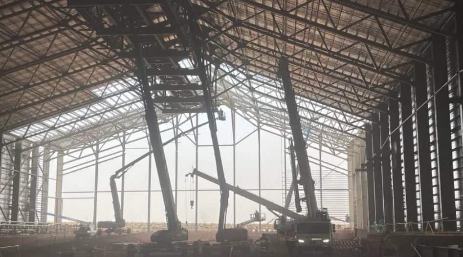 Several cranes and construction equipment operate inside a large, partially built warehouse with a high, metal frame roof. Sunlight streams through the open wall panels, illuminating the structural beams and machinery.