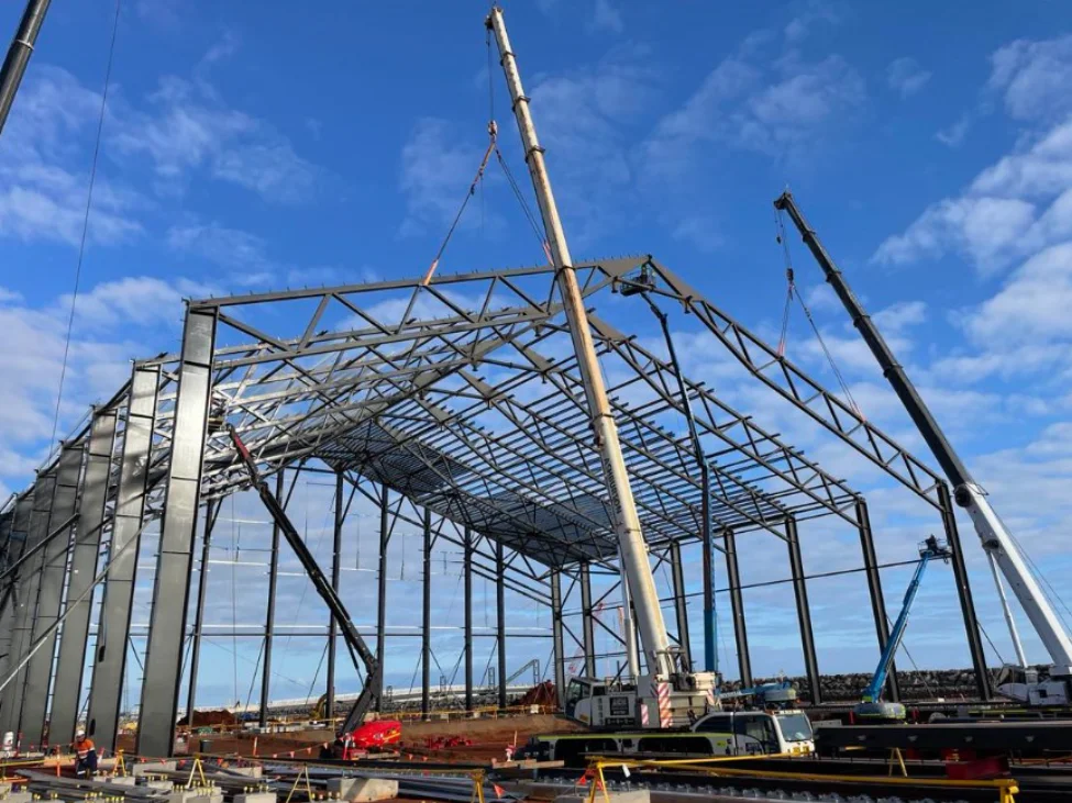 Cranes and construction workers assemble the steel framework of a large industrial building under a blue sky with scattered clouds. Construction materials and equipment are visible on the site.