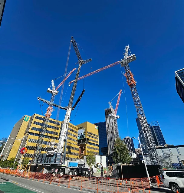 Several large construction cranes operate at a building site in a city, surrounded by tall office buildings under a clear blue sky. Orange safety barriers line the street in the foreground.