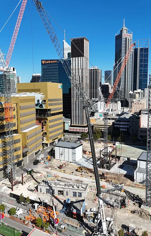 A city construction site with several cranes, surrounded by tall office buildings under a clear blue sky. Construction equipment and materials are scattered on the ground.