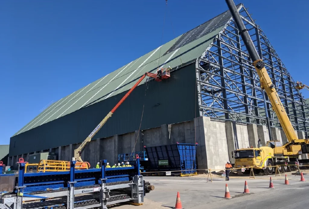 A large industrial building under construction, with workers on elevated platforms, a yellow crane, construction equipment, and safety cones in the foreground under a clear blue sky.