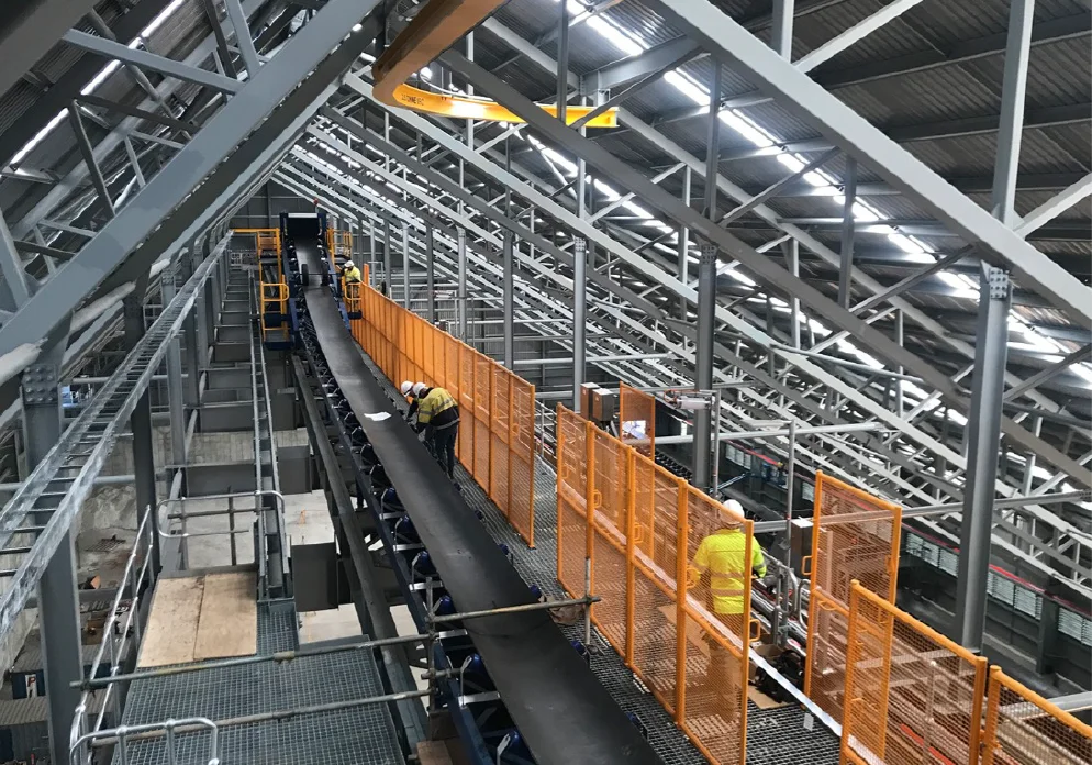 Workers wearing safety gear inspect and work on a long conveyor belt system inside a large industrial building with metal beams and orange safety fences.