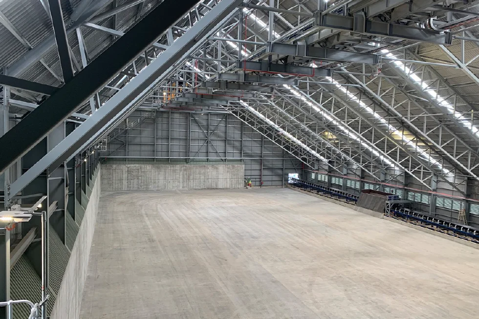 Large, empty industrial warehouse with a high, sloped metal roof, exposed beams, and concrete floor and walls. Two small figures stand near the far wall, emphasizing the scale of the space.