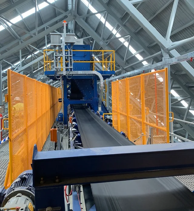 A black conveyor belt runs through a large industrial facility with metal beams, orange safety railings, and machinery under a high, sloped roof.