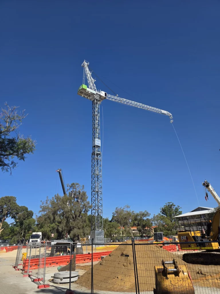 A tall construction crane stands at a building site on a clear day. The area is fenced off with orange barriers, and construction vehicles and equipment are visible on the ground. Trees and buildings are in the background.