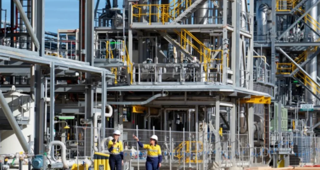 Two workers in safety gear walk and talk in front of a large industrial plant structure with metal pipes, staircases, and platforms. The facility appears complex with yellow railings and various equipment.