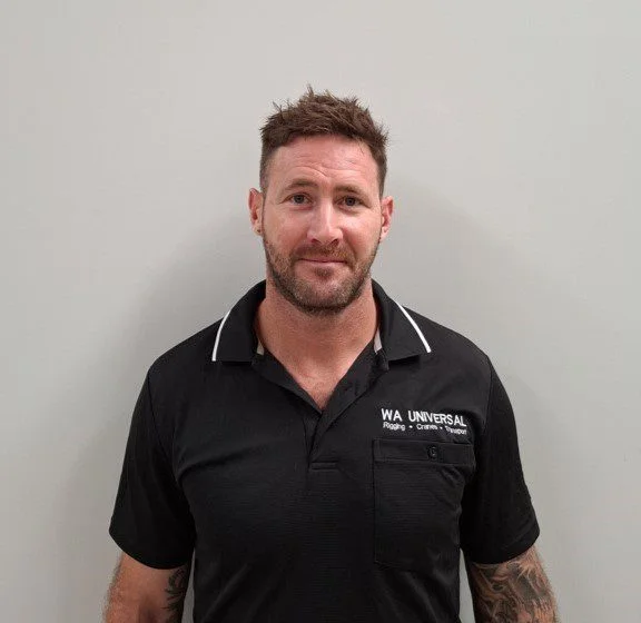 A man with short brown hair and a trimmed beard stands against a plain light-colored wall, wearing a black collared shirt with "WA UNIVERSAL" and other text on the chest. He has visible tattoos on his arms.