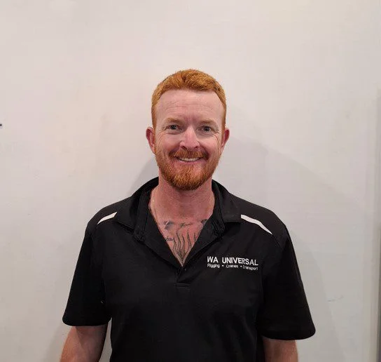 A man with short red hair and a beard is standing and smiling. He is wearing a black collared shirt with "WA Universal" and other text on the chest. The background is a plain, light-colored wall.