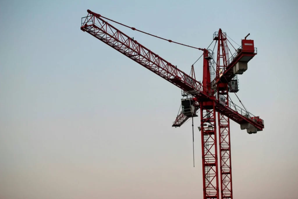A large red construction crane stands tall against a clear sky at dusk, with its long arm extending horizontally and several platforms and cables visible.