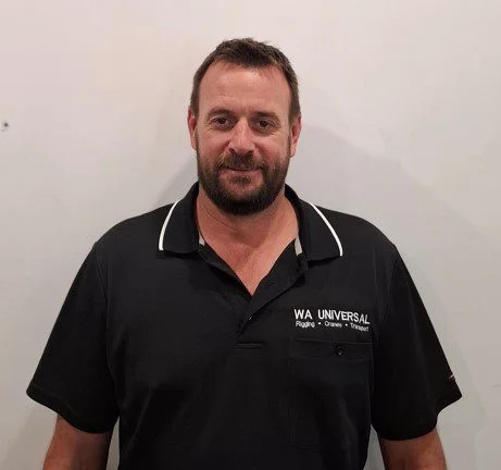 A man with short brown hair and a beard stands against a plain white wall, wearing a black polo shirt with "WA UNIVERSAL" written on the chest.