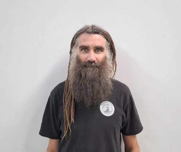 A person with long brown dreadlocks and a full, bushy beard stands against a plain white background, wearing a black t-shirt with a round logo on the left side.