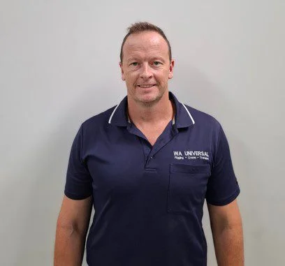 A man stands against a plain light-colored wall, wearing a navy blue polo shirt with "WA UNIVERSAL" written on the chest. He is smiling and looking directly at the camera.