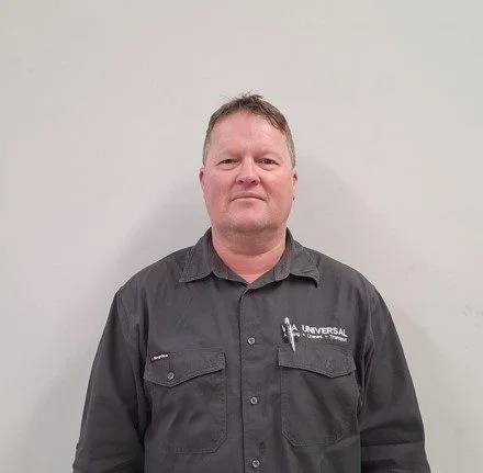 A man with short hair wearing a dark grey button-up shirt with "UNIVERSAL" and a small logo on it stands against a plain, light-colored wall.