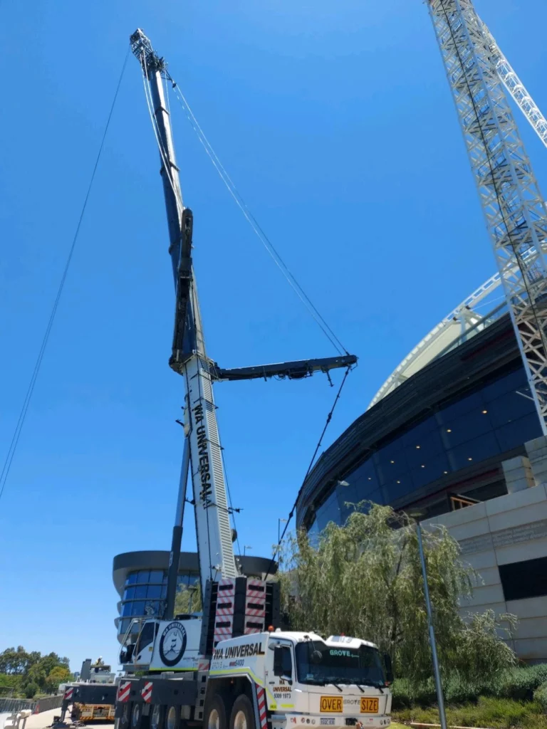 A large construction crane from "THE UNIVERSAL" lifts materials beside a modern stadium building under a clear blue sky, with trees and another crane visible in the background.
