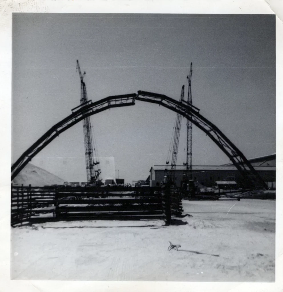 Black and white photo of a large, incomplete metal arch under construction, with cranes on each side and industrial buildings in the background. The foreground shows a fenced area and a dirt ground.