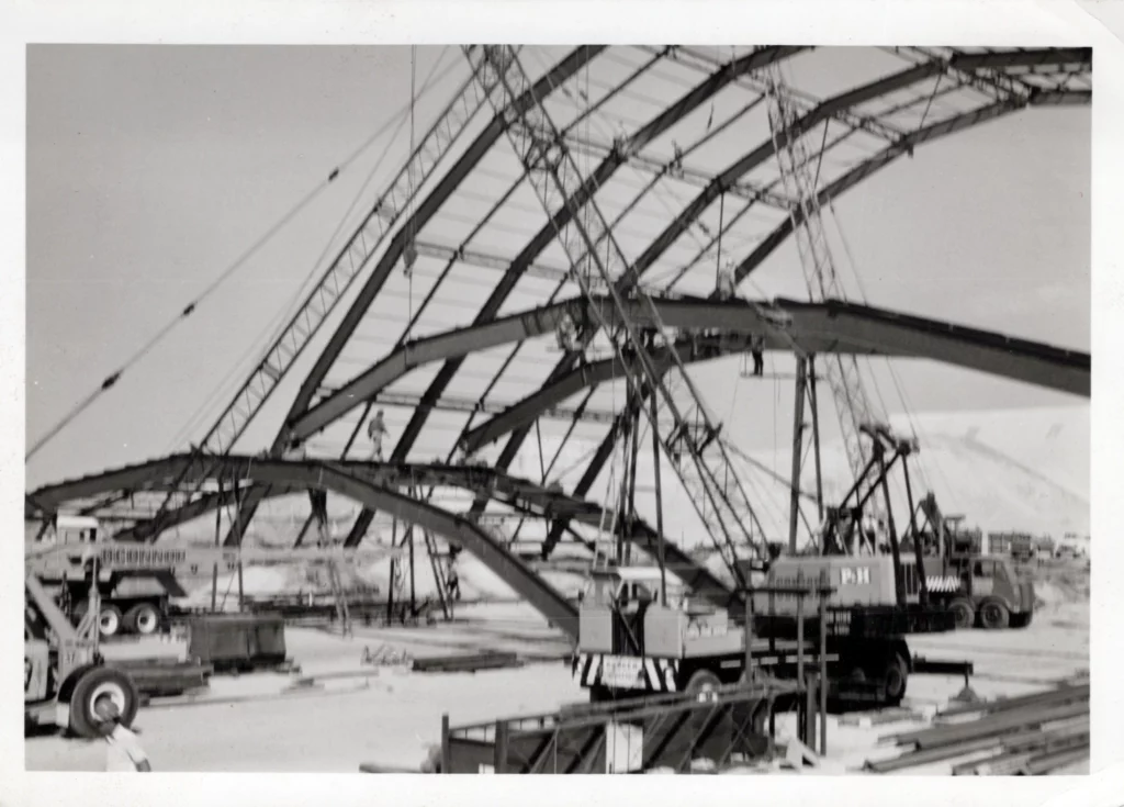 Black and white photo of a large, arched steel structure under construction, with cranes, workers, and construction equipment visible on the site. The framework forms several curved beams.