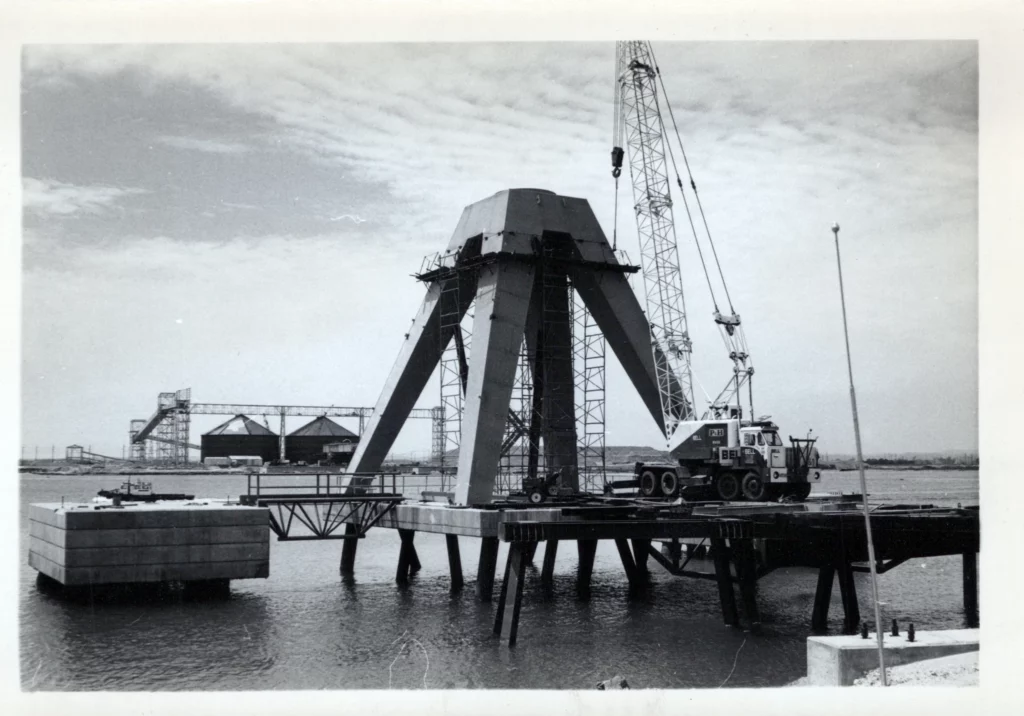 Black and white photo of a large crane positioned on a dock, lifting a heavy, four-legged industrial structure over water. Additional structures are visible in the background under a partly cloudy sky.