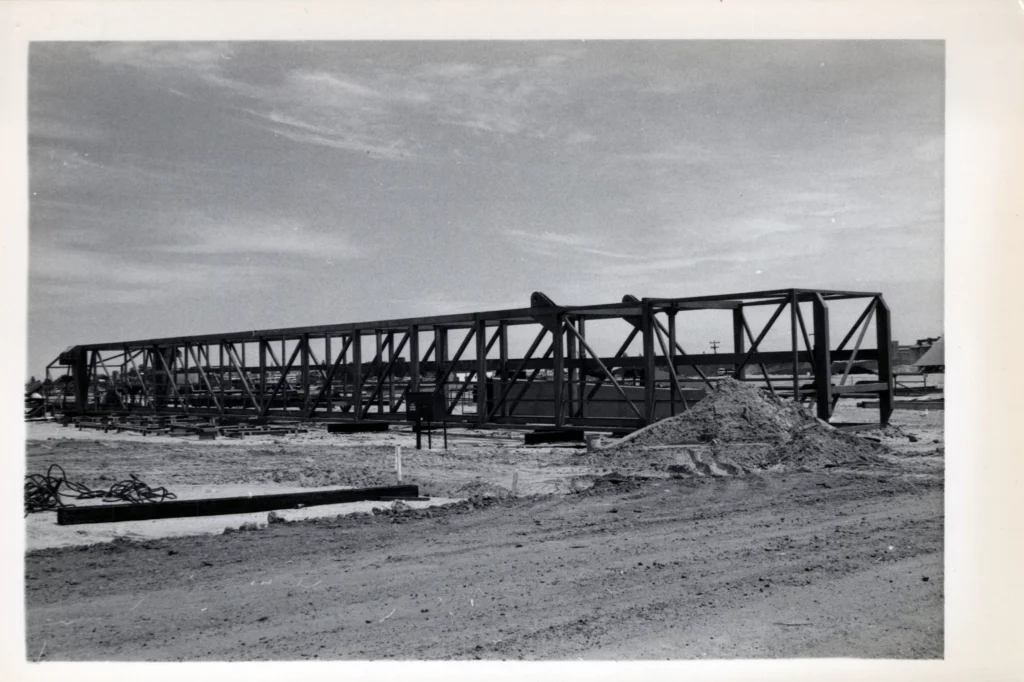 A large steel truss structure is under construction on a dirt lot, with piles of sand and scattered building materials in the foreground, under a partly cloudy sky.