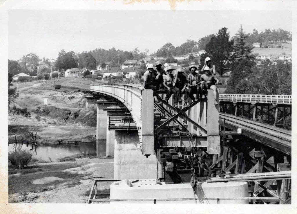 Black and white photo of six construction workers sitting on steel beams of a partially built bridge over a river, with a rural town and trees visible in the background.