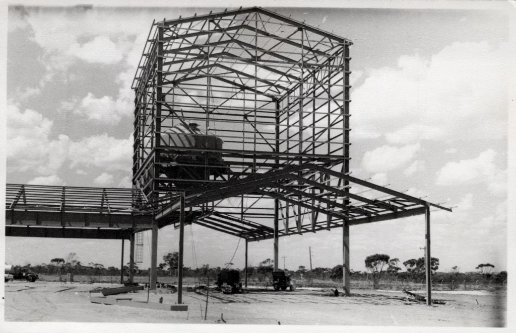 A large steel frame structure under construction stands elevated on pillars, with beams and girders exposed. Several vehicles and trees are visible in the background under a partly cloudy sky.