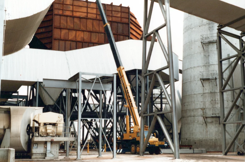 A large yellow crane is positioned at an industrial site, surrounded by metal structures, a concrete tower, and corrugated metal roofing, with a rust-colored building section in the background.