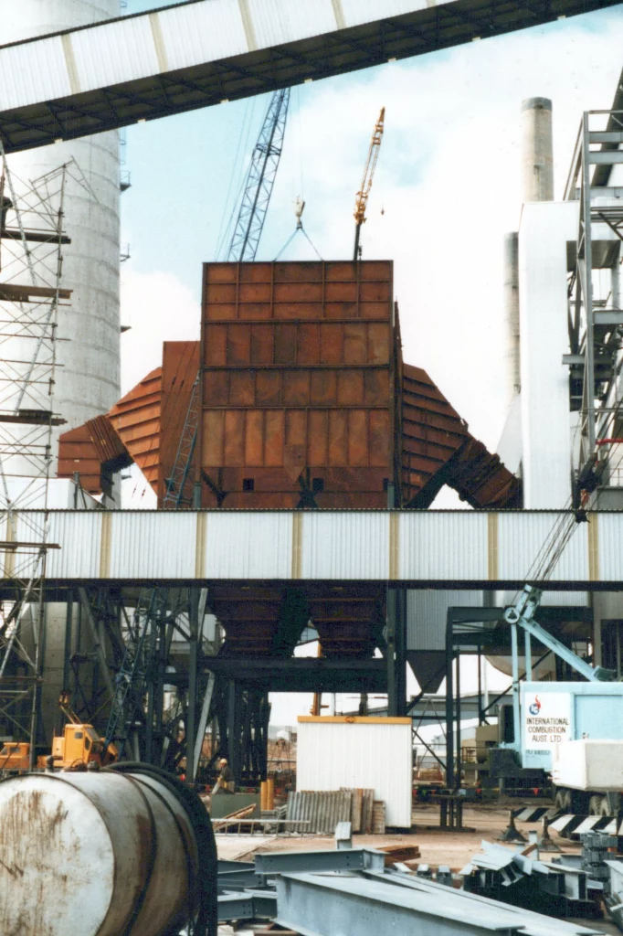 A large industrial structure with a rust-colored central unit, two cranes, scaffolding, and construction equipment surrounding it at an outdoor construction site.