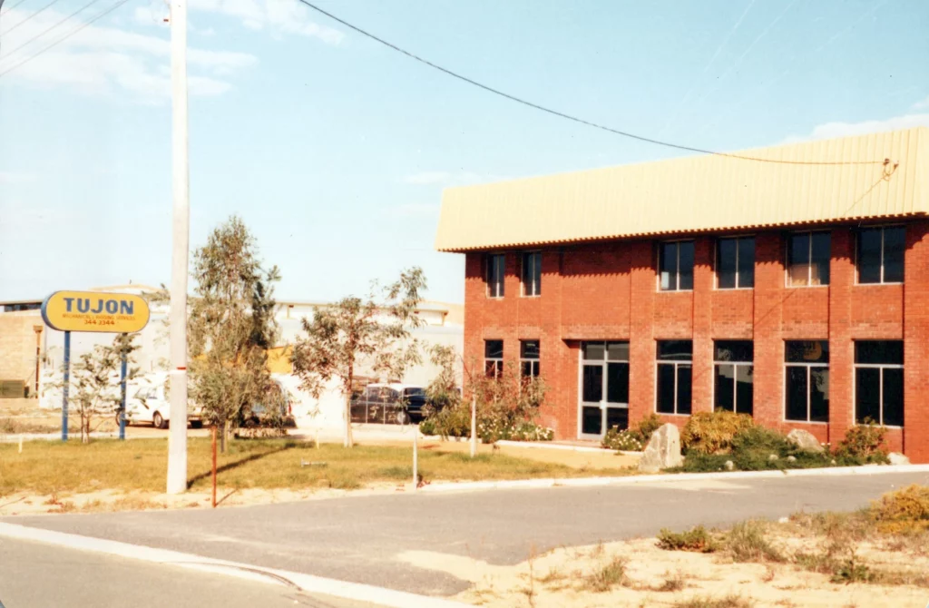 A two-story red brick building with large windows sits on a corner lot. A yellow sign reading "TUJON" is visible on the grass near the sidewalk, with some trees and a few parked cars nearby.