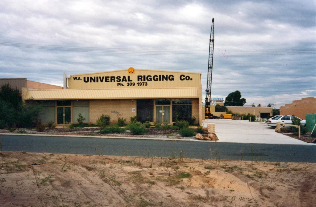 A tan building with a sign reading "W.A. Universal Rigging Co. Ph. 309 1973" sits under a cloudy sky. A crane stands in the lot, and a green car is parked near the building. Shrubs and dry grass line the road.